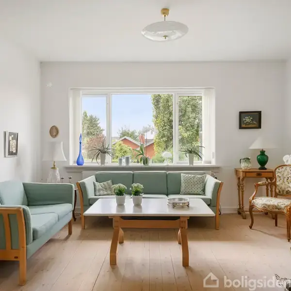Stue med lyseblå sofa og sofabord i træ, omgivet af grønne planter. Store vinduer slipper naturligt lys ind og viser udsigt til have med træer. Elegant, klassisk indretning.