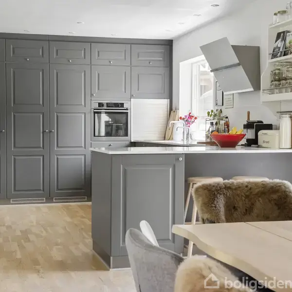 Gråt, moderne køkken med indbyggede skabe og ovn. En central køkkenø har en rød skål og planter. Spisebordsstole har pelsbetræk. Lyse trægulve og væggehængte hylder med planter.