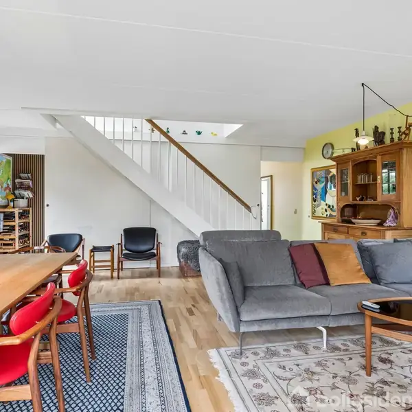 Gråt sofaarrangement med puder står ved et sofabord, omgivet af en træreol og farverige stole ved et spisebord. Baggrunden har en gul væg og en trappe til overetagen.