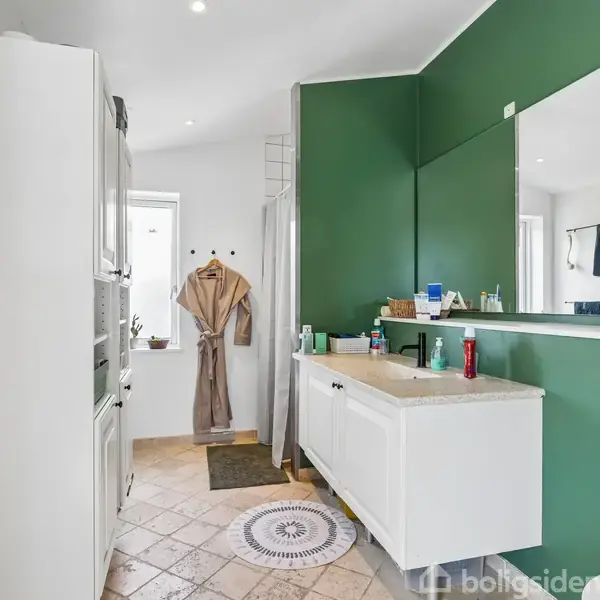 Et badeværelse med grønne vægge har et hvidt skab og håndvask. Hylden er fyldt med toiletsager. Et brusebad står ved siden af en ophængt morgenkåbe. Gulvet har beige fliser.