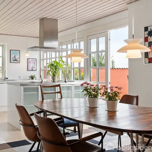 Spisebord med stole omgivet af moderne køkken. Planter pynter bordet, mens store vinduer lader lys ind. Køkkenet har hvidt interiør og vægdekorationer. Spiseområdet har hængende lamper.