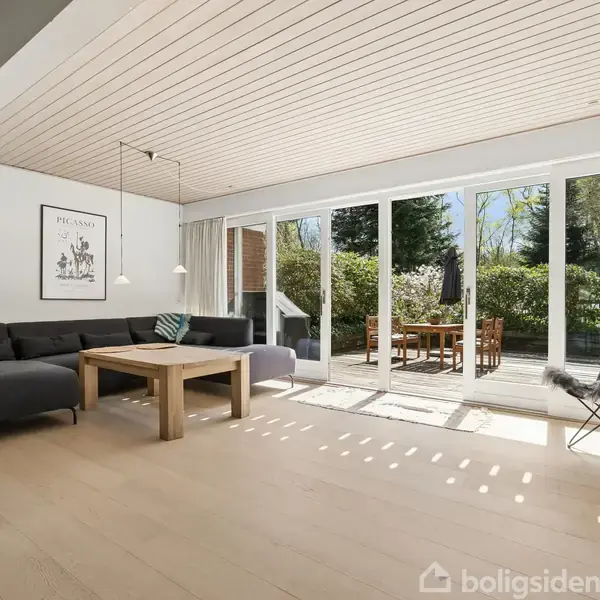 Stue med sort sofa og træbord, sollys strømmer gennem store glasdøre, åbner op til terrasse med havemøbler. Vægplakat over sofa, gulvlampe til venstre. Afslappet, lys atmosfære.