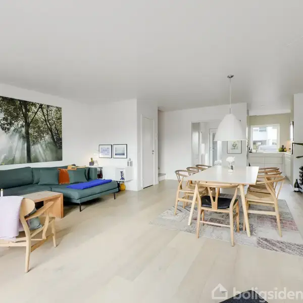 Stue med en grøn sofa og træmøbler arrangeret omkring et sofabord. Et stort naturbillede hænger på væggen, og et spisebord med seks stole ses i baggrunden. Ingen tekst fundet.