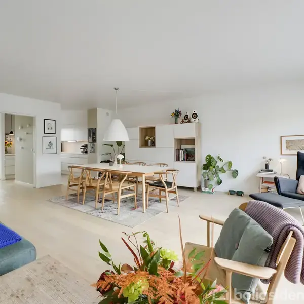 En lys, moderne stue indeholder spisebord med stole i midten, omgivet af reoler og planter. En lænestol med tæpper står ved et stort vindue, hvilket skaber en indbydende atmosfære.