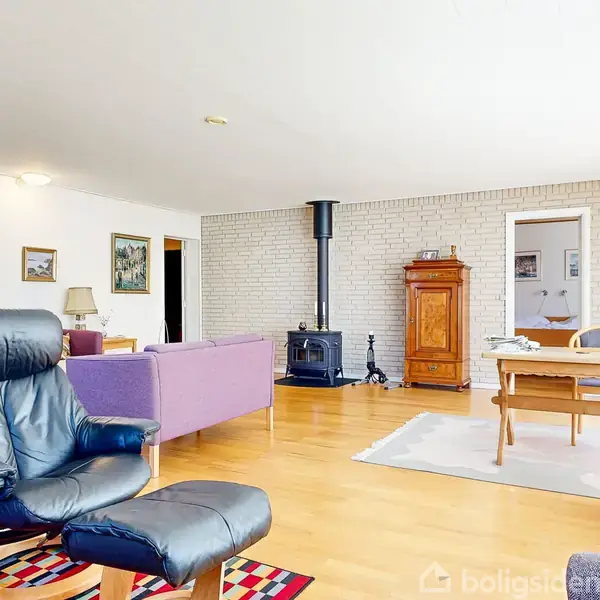 Sort læderlænestol står foran farverigt tæppe. Stuen er lys med trægulve, pejs, lilla sofa, træmøbler og hvide vægge dekoreret med malerier.