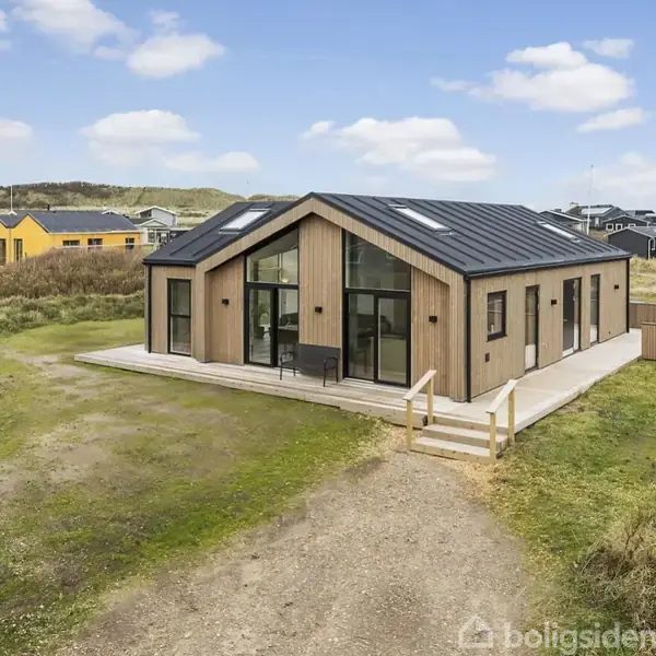 Træhus med store vinduer står på en græsbelagt grund, omgivet af andre huse. Det har en sort tagkonstruktion og terrasse, samt en udendørs jacuzzi i nærheden.