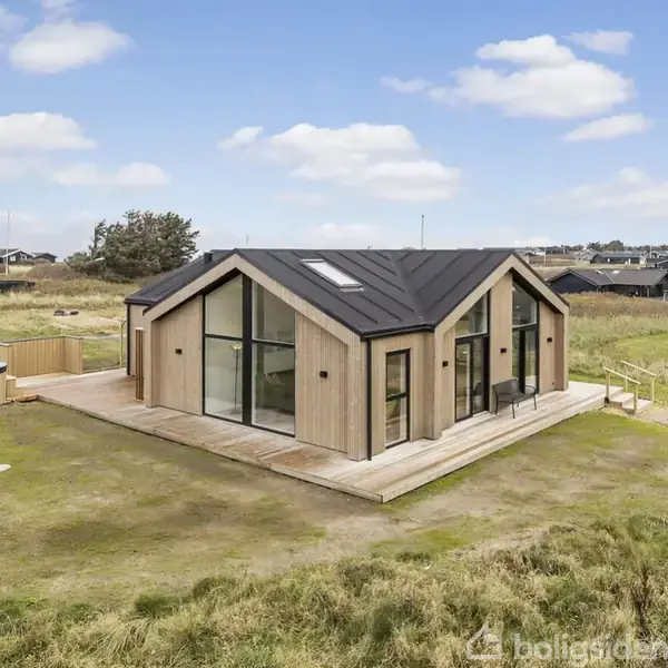 Et moderne træhus med store glaspartier står på en græsbevokset grund i et kystnært område. Flere lignende huse ses i baggrunden under en skyfri himmel.