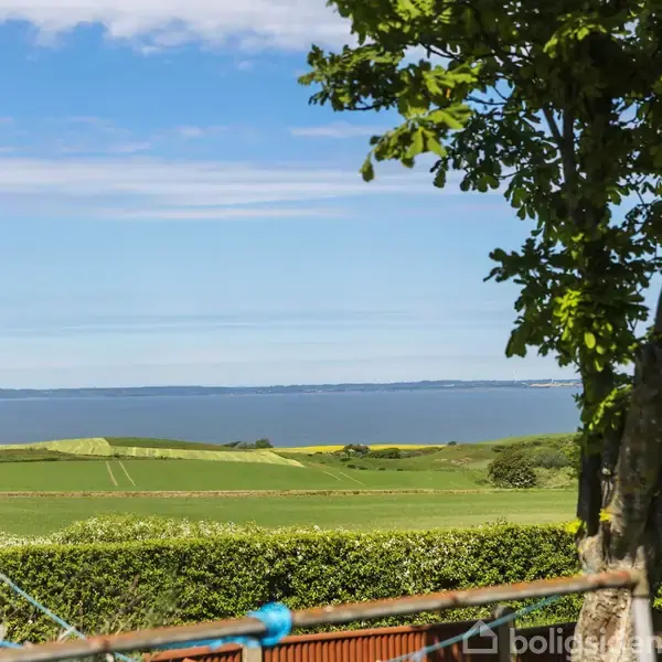En vindmølle står stille på en frodig, grøn mark ved kysten, mens en blå himmel med lette skyer strækker sig over horisonten; et træramme i forgrunden.
