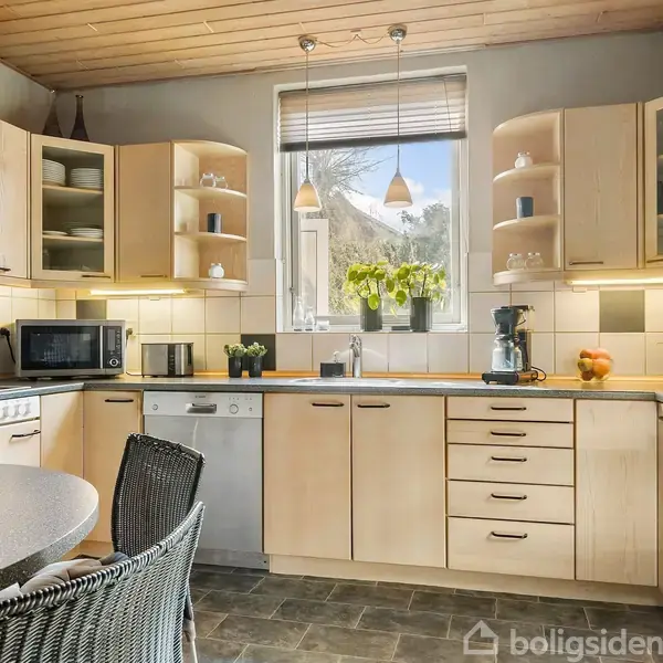 Et lyst køkken med træskabe omgiver et rundt spisebord. En mikrobølgeovn står på bordet, og en kaffemaskine ses nær vinduet, der giver udsigt til natur.