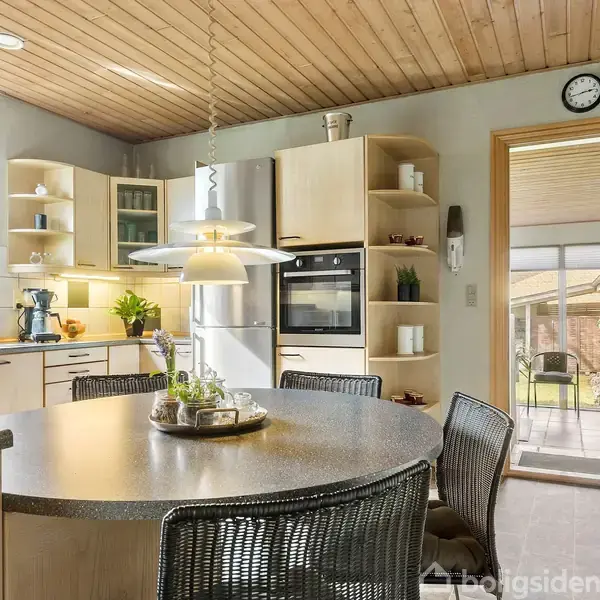 Et lyst køkken med runde spisebord omkranset af stole. Køleskab og ovn indbygget i trækøkkenelementer. Store vinduer, planter, og udsigt til terrasse giver rummet et åbent, venligt indtryk.