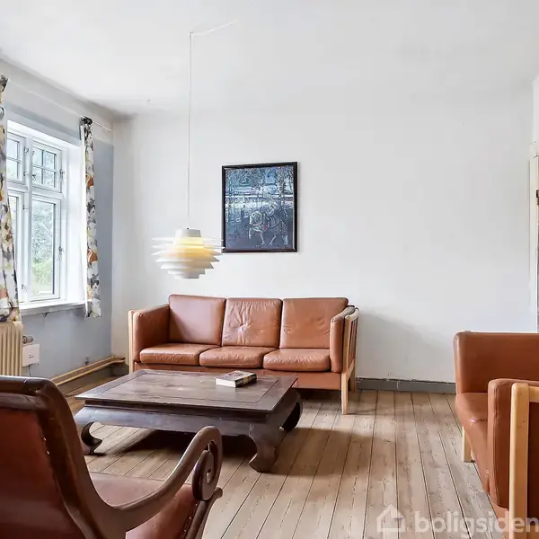 Stue med lædersofa og to lænestole i brun, stående omkring et træbord. Lampe hænger over bordet. Væggen har kunst, og der er et vindue med blomstrede gardiner, som lukker lys ind.