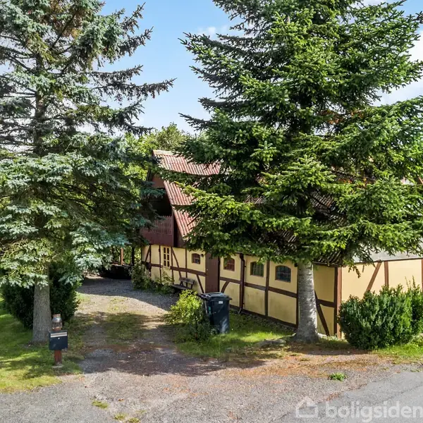 Bindingsværkshus med rødt tegltag står blandt høje grantræer. Grusvej foran huset leder til grønne arealer og marker i baggrunden. En postkasse bemærkes i forhaven.