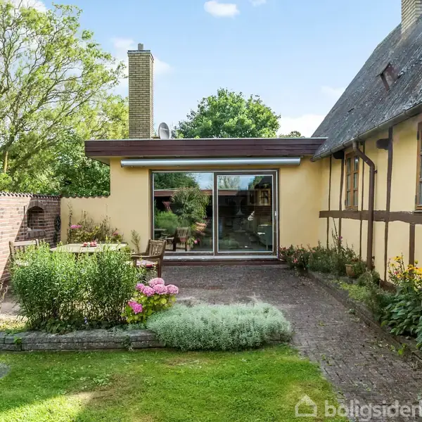 Et gult hus med stråtag har en stor glasdør mod en brostensbelagt terrasse. Blomsterbede omkranser terrassen, og en murstensmur skaber afskærmning i haven.