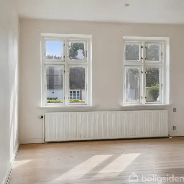 Et lyst rum med trægulv, hvor to vinduer med hvid ramme skaber en lys atmosfære. Udenfor ses et hustag og træer. Der er en radiator under vinduerne.