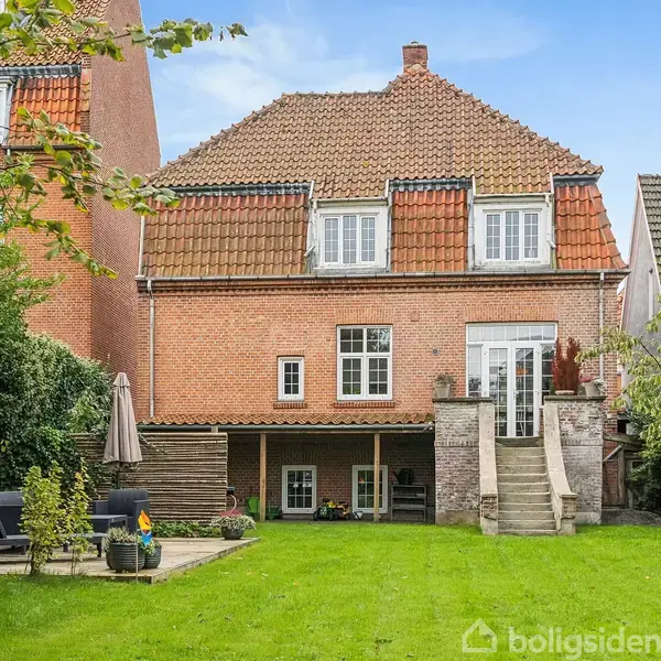 Rødstenshus står stille i en frodig have. Husets tage er røde og har flere vinduer. Haven er velplejet med en terrasse og siddeområde til venstre med havemøbler.