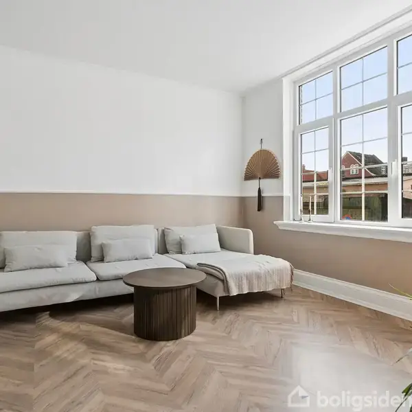En lys stue med en beige sofa placeret mod en væg. Et rundt sofabord foran sofaen. Store vinduer viser udsigt til huse udenfor. Gulvet er i sildebensparket.