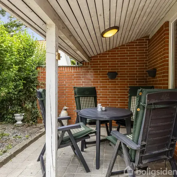 Et rundt bord med fire stole står under en overdækket terrasse. Møblerne er placeret ved en murstensvæg, omgivet af grønne planter og belagt med fliser.