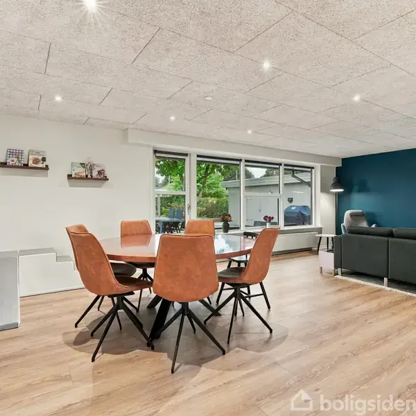 Rundt spisebord med seks brune stole står i en lys stue. Store vinduer giver udsigt til en grøn have. En grå sofa står ved en blå væg.