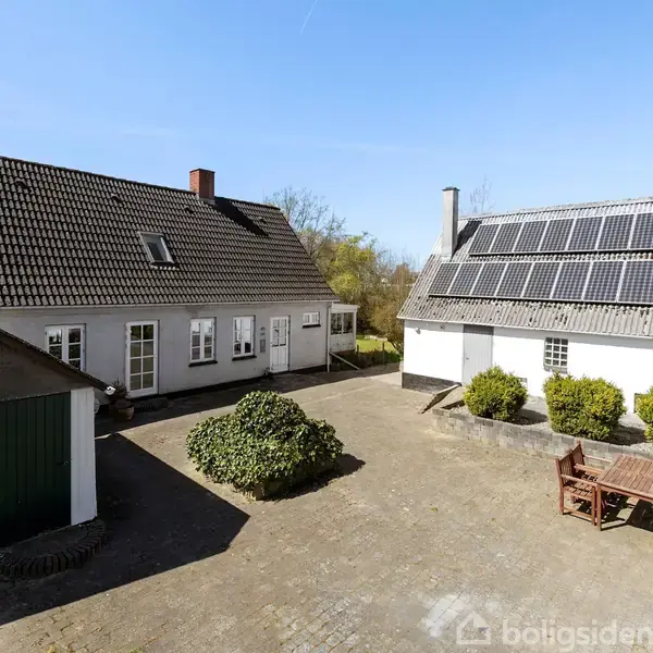To bygninger står overfor hinanden i en gårdsplads med stenbelægning. Den ene bygning har solpaneler på taget. En træbænk med bord står til højre.