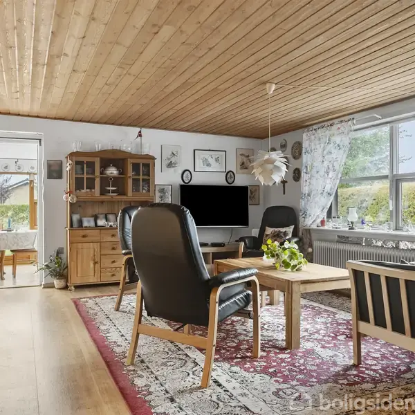 En stue med lænestole og en sofa omkring et sofabord, på et orientalsk tæppe. Et vitrineskab står ved væggen. Store vinduer giver udsigt til haven udenfor.