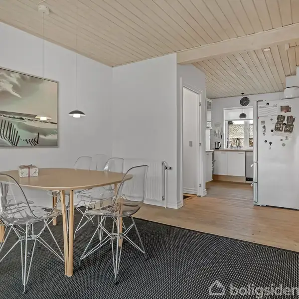Et spisebord med gennemsigtige stole står på et tæppe i en spisestue. Billedramme på væggen. Åben adgang til køkken med hvid køleskab, trægulve, og hvidt loft.