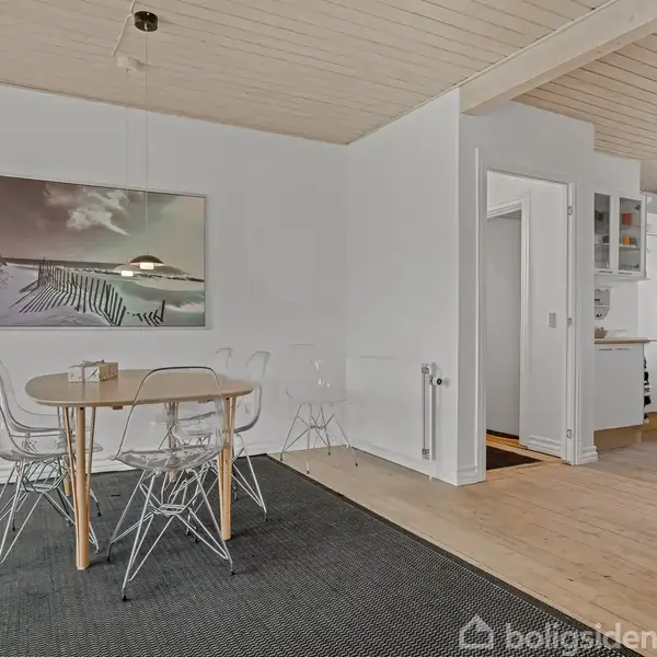 Rundt træbord med gennemsigtige stole står på et sort tæppe. Baggrunden har hvidmalede vægge og et billede med strandmotiv. Til højre ses en køkkenindgang med hvidt køleskab.