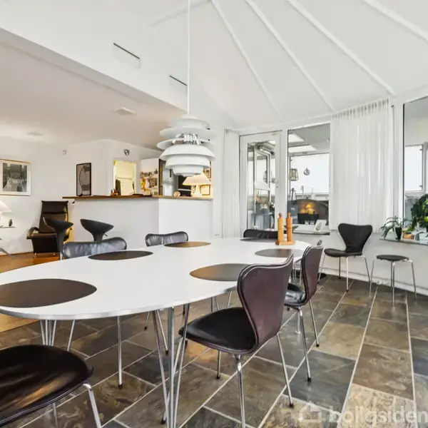 Et ovalt spisebord med mørke stole står i et lyst, flisebelagt rum. Store vinduer og planter skaber en moderne og lys atmosfære. En lampe hænger over bordet.