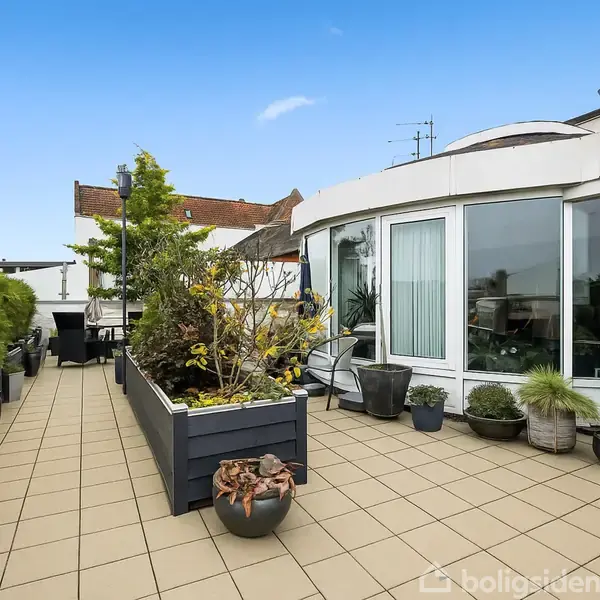 Terrasse med fliser og krukker med planter placeret langs kanterne. En buet glasfacade i baggrunden viser døre og vinduer. Skylinen viser murstensbygninger og skorstene i det fjerne.
