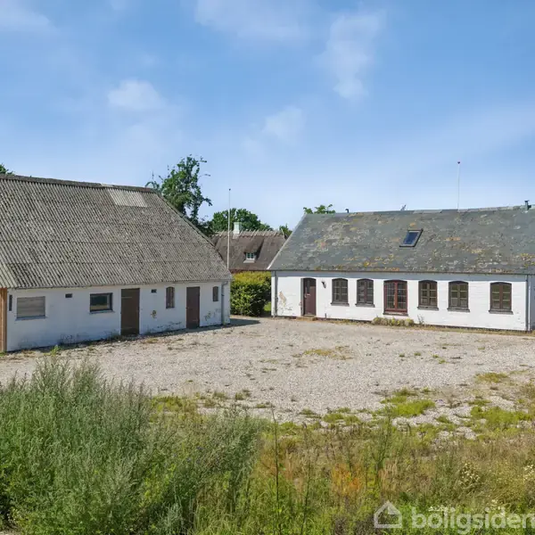 To bygninger står i en grusbelagt gårdsplads omgivet af grønt landskab. Den ene bygning har et metal- og trætag, mens den anden har skrå tage med flere ovenlysvinduer.