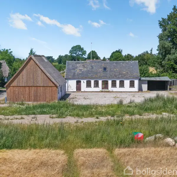 En hvid bygning med gråt tag ligger ved siden af en trælade. Foran er en blanding af græs og mark med buske og træer i baggrunden under en blå himmel.