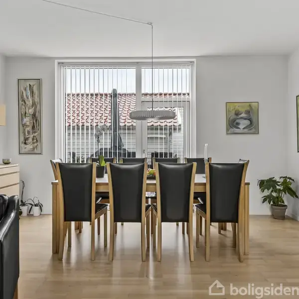 Spisebord med seks stole står i en lys spisestue. En stor vinduesparti med persienner giver udsigt til hustage. Væggene har malerier, og der er en kommode til venstre.