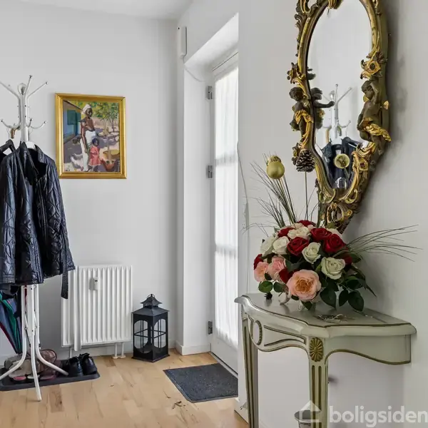 Et smalt gangområde med en hvid væg er dekoreret med et gyldent spejl og et bord med blomster. Jakker hænger på en knagerække, og en lanterne står ved en radiator.