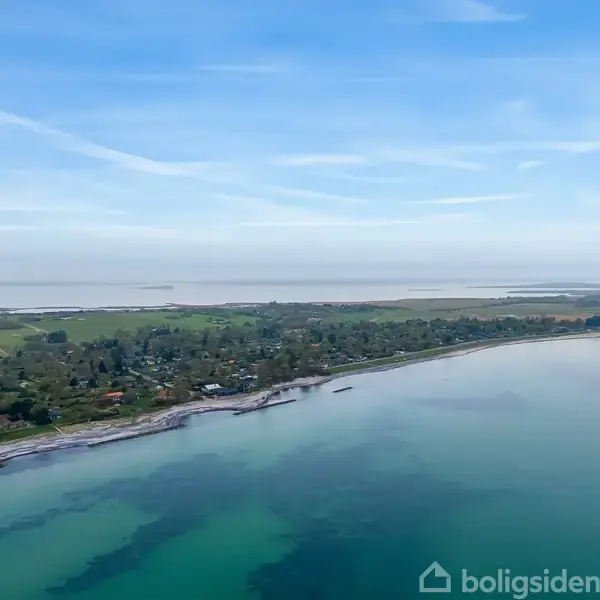 Kyststrækning strækker sig langs en rolig havoverflade; grønne marker og spredte huse danner baggrunden, mens himlen er oversået med lette skyer.