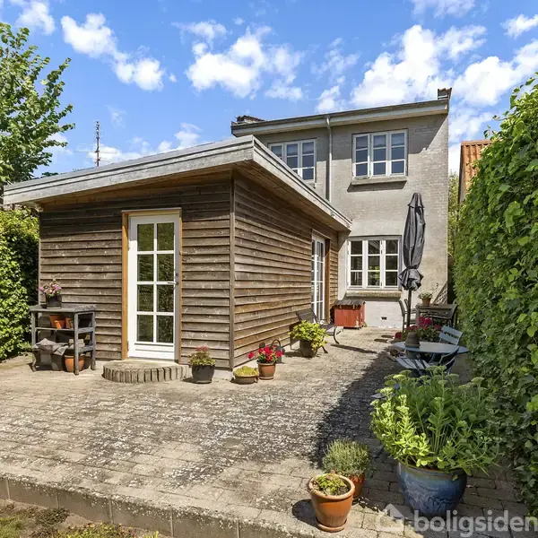 Træbeklædt skur står i en gårdhave med flisebelægning, omgivet af grønne hække. Ved siden af er et større hus med et havebord og parasol. Blomsterkrukker dekorerer området.