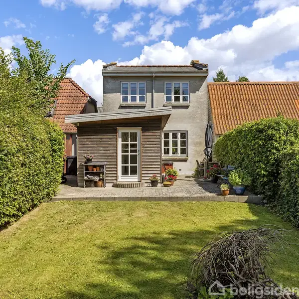 Et gråt hus med rødt tegltag står i en have omkranset af hække. Der er terrasse med blomsterkrukker foran huset under en blå skyet himmel.
