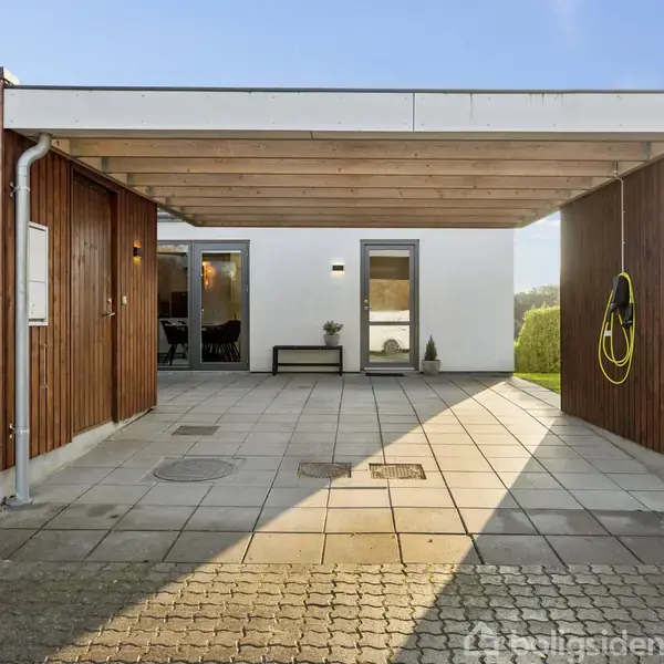 Carporten er åben, træbeklædt med fladt tag, hvor en gul haveslange hænger på væggen. To firkantede vinduer ses på husets facade, flisebelagt gulv. Adressen er "Ahornvænget 11".