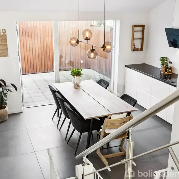 Et spisebord omgivet af sorte stole står i et moderne køkken-alrum med lyse vægge. Over bordet hænger en stilfuld lampe. I baggrunden ses en stor glasdør til en terrasse.