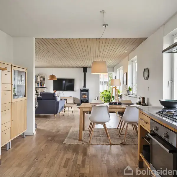 Køkkenalrum med spisebord, omgivet af træmøbler og hvide stole, i et lyst rum med store vinduer og moderne indretning. Køkkenet har gasblus, og stuen har sofa og tv.