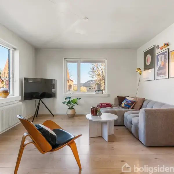 Stue med grå sofa og lavt, rundt bord placeret på lakeret trægulv. TV på stativ står ved vinduer med udsigt. Kunstplakater dekorerer den lyse væg. Grøn plante i hjørnet.