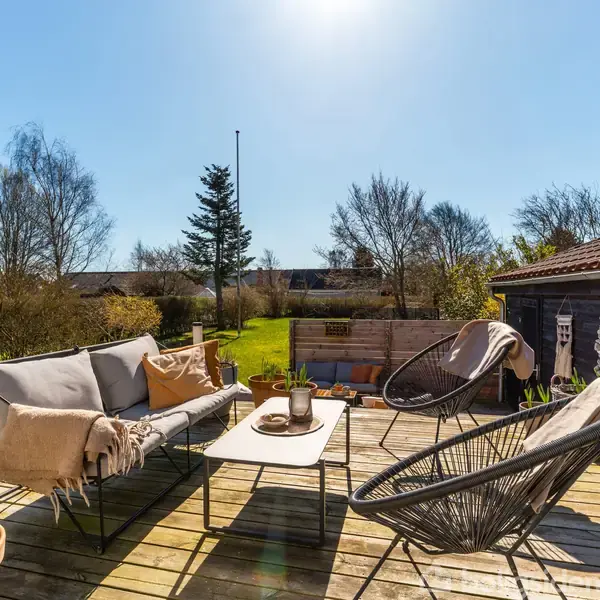 Terrassemøbler står roligt på et trædæk, omringet af planter og græsplæne. Møblerne omfatter en sofa, to stole og et bord. Sollys skinner overalt i en baghave omgivet af træer.