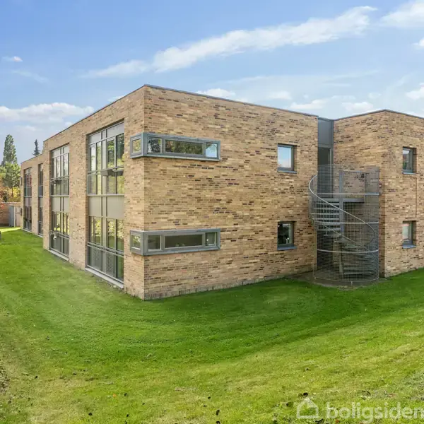 Murstensbygning med to etager, spiraltrappe på siden, placeret på en grøn plæne. I baggrunden er der træer i efterårsfarver og en parkeret bil til højre.