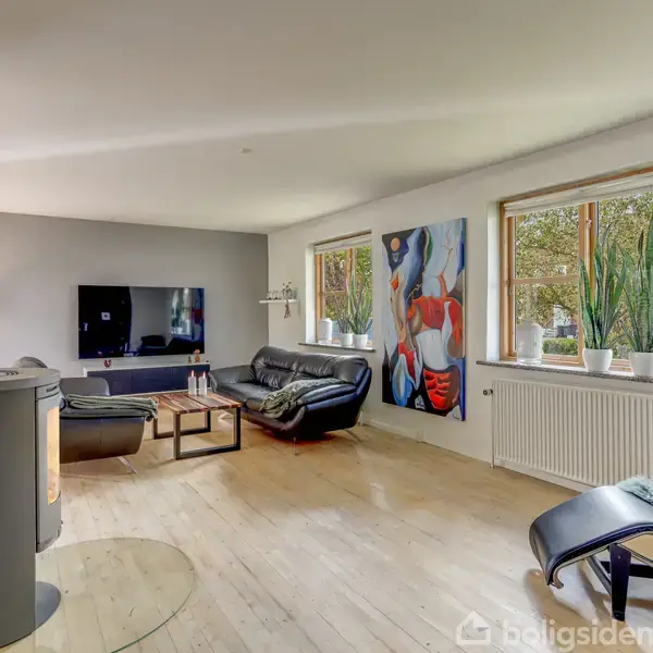 Stue med sorte lædersofaer og hvilestol foran et vægmonteret TV. Store billeder pryder væggen, og planter står på radiatoren under store vinduer. En rund brændeovn ses i forgrunden.