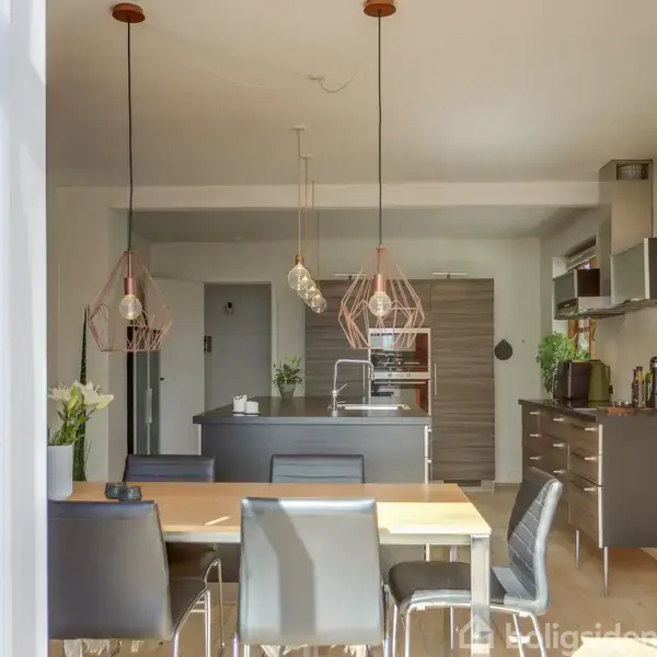 Spisebord med seks stole står i et moderne køkken. Loftslamper hænger over bordet. Køkkenet har mørke skabe, rustfri apparater og planter, i et rum med lysindfald.