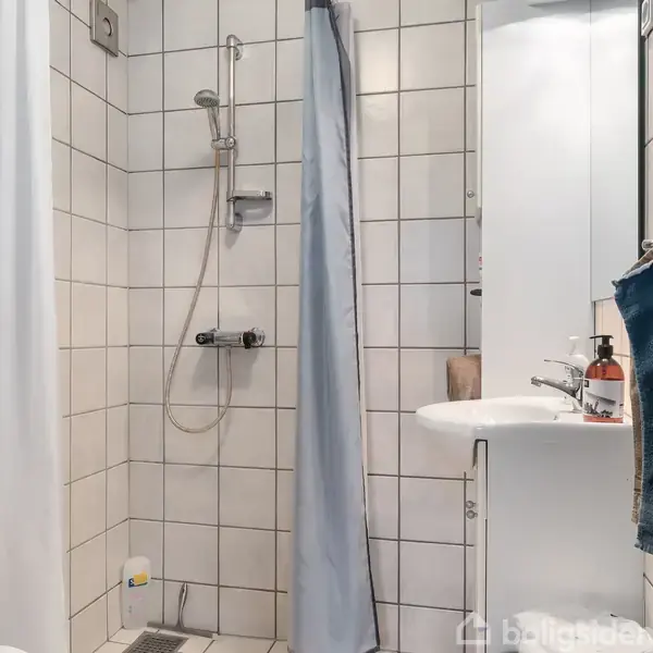 Brusekabine med en regnbruservej hænger på væggen, omgivet af lyse fliser og et hvidt bruseforhæng. Ved siden af er en vask med spejl og toiletpapirholder.