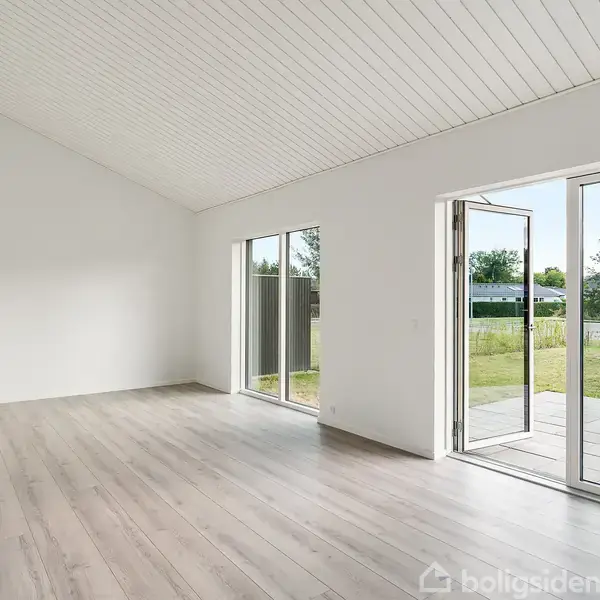 Et lyst rum med trægulv og hvide lofter har dobbeltdøre, der åbner ud til en terrasse. Udenfor ses en græsplæne og træer i baggrunden. Ingen tekst til stede.