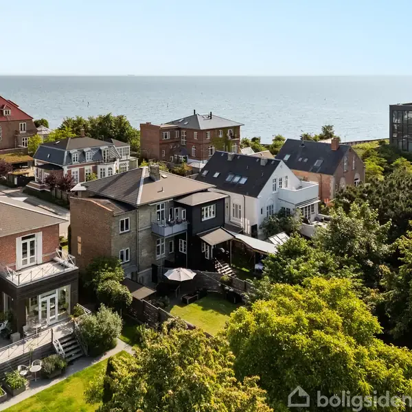 Flere huse ligger langs en kystlinje med udsigt til havet. Bygningerne har store terrasser og grønne haver, omgivet af træer og tæt bebyggelse.