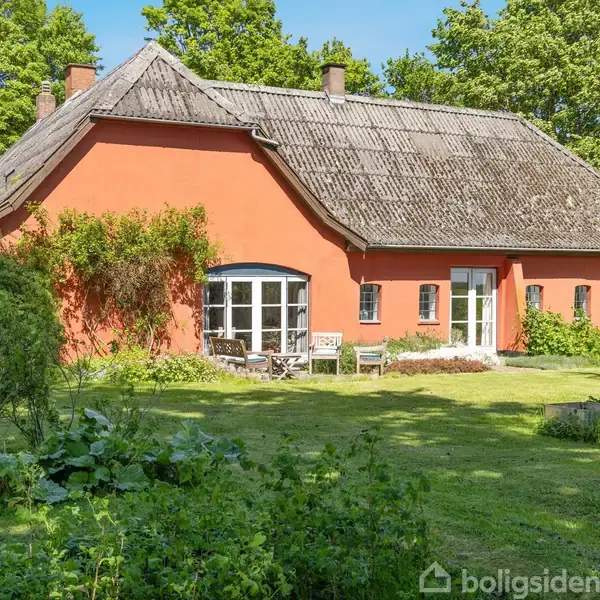 Et rødt hus med skråt tag, omgivet af grønne træer og en frodig have. Der står havemøbler foran huset ved en stor glasdør.