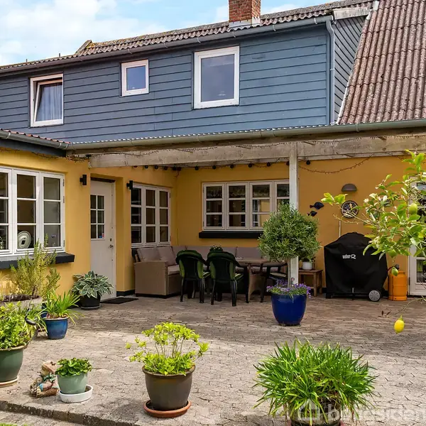 Gul murstenshus med blå overetage står i en gårdhave. Der er mange potteplanter, bord med stole, og en grill. Området er flisebelagt og ser fredfyldt ud.