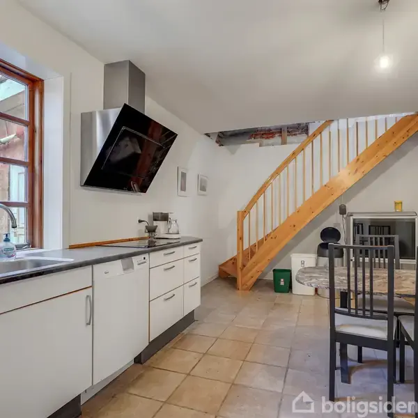 Et lyst køkken med hvide skabe. En trappe fører til en anden etage. Et spisebord med stole er til stede. Store vinduer giver udsigt til en rød bygning udenfor.