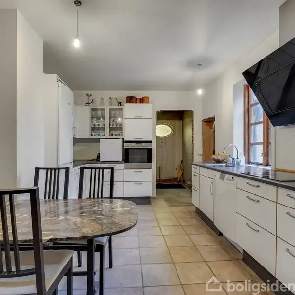 Et køkken med et rundt marmorbord og fire stole. Der er hvide skabe og en sort emhætte. Rummet er lyst med flisegulv og vindue til højre.